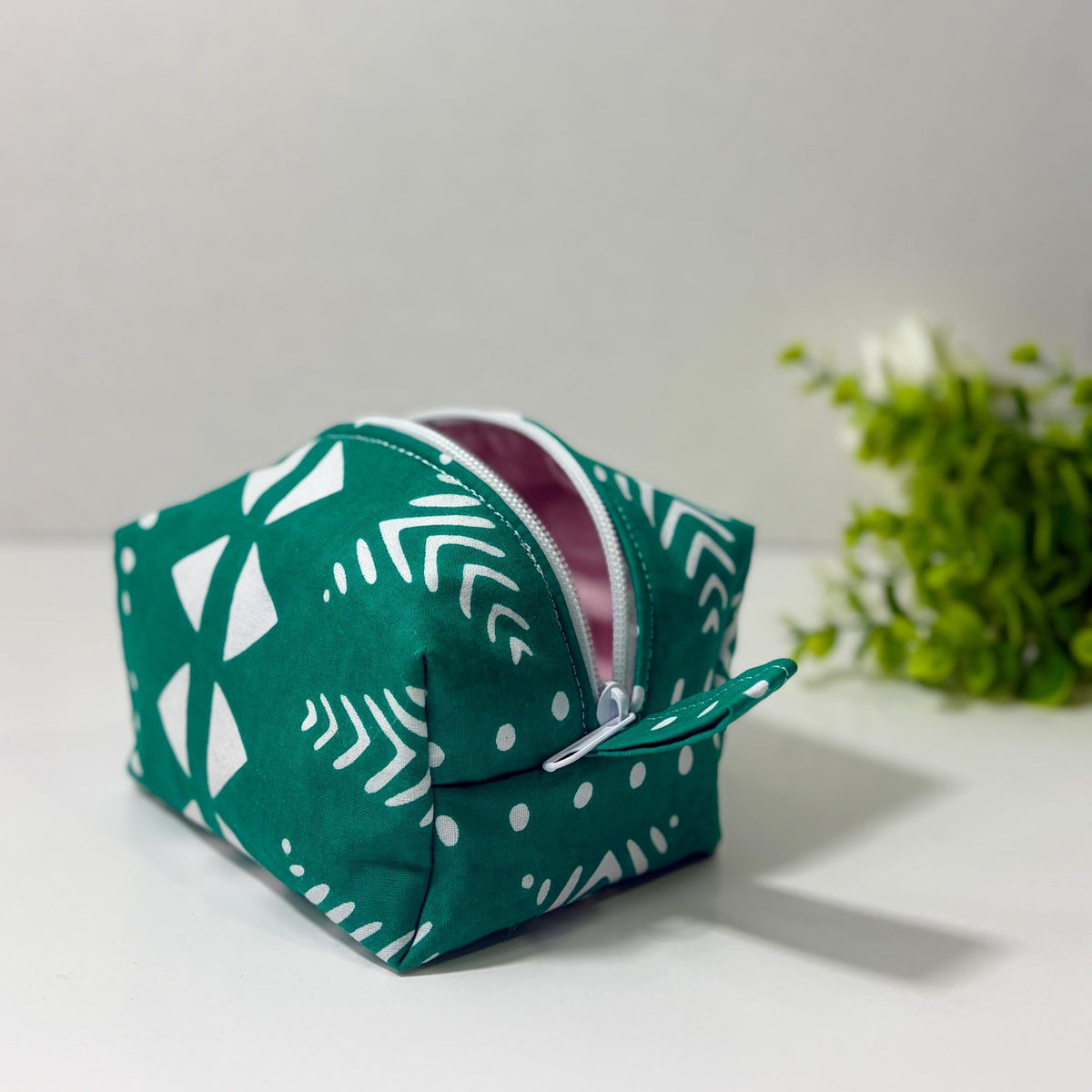 A green and white patterned cosmetic bag with a zipper closure is placed on a white surface, with a plant in the background.
