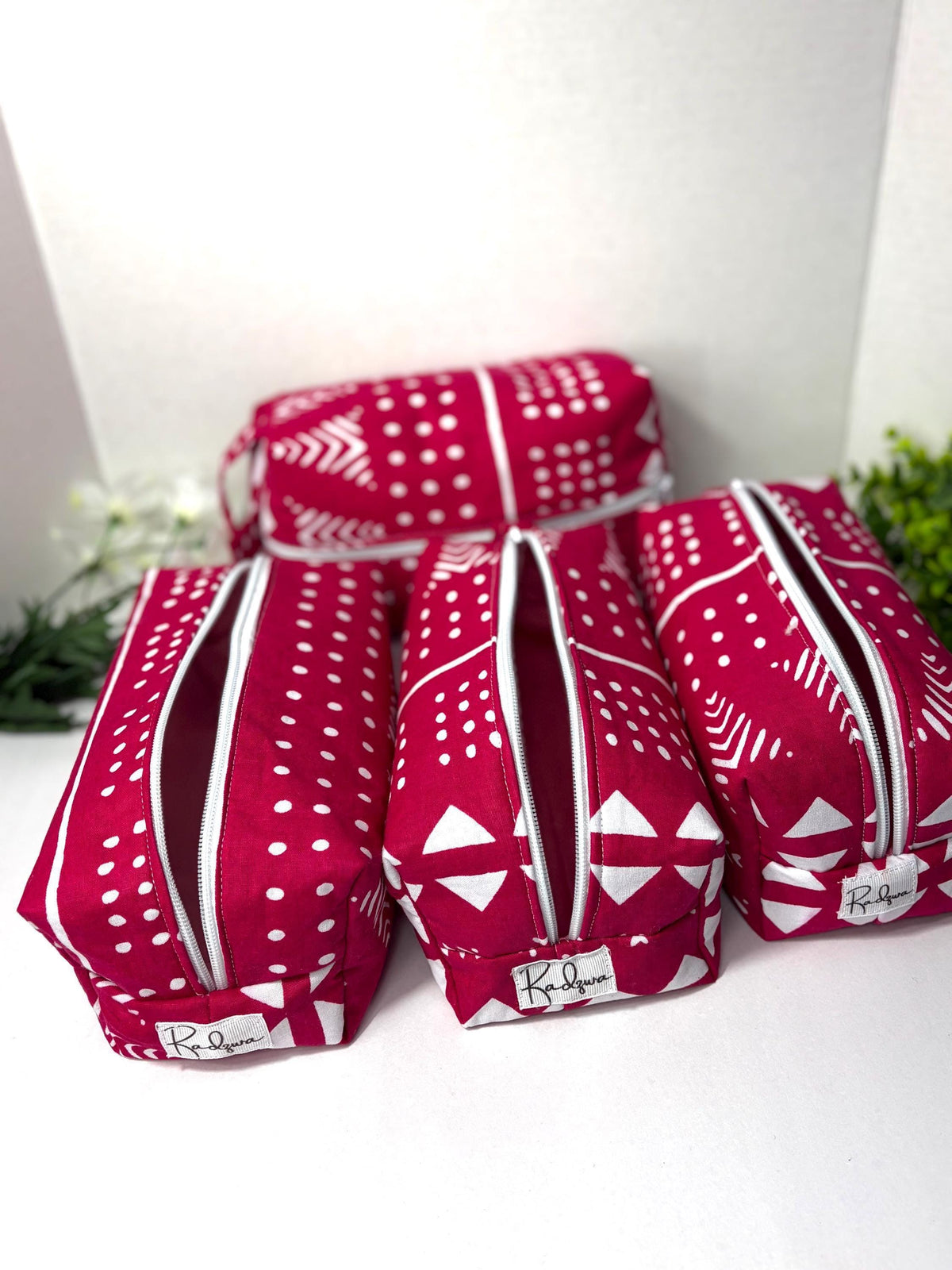 Three red and white patterned bags with zippers are arranged on a white surface, with a plant visible in the background.