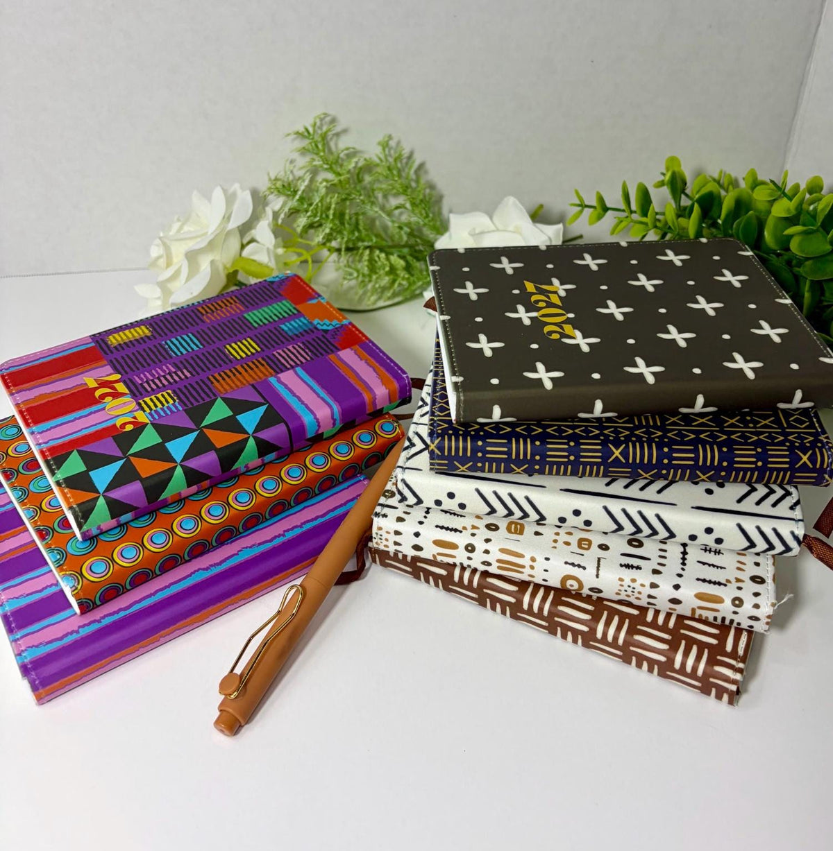 A stack of colorful patterned notebooks and a pen on a white surface, with some greenery in the background.