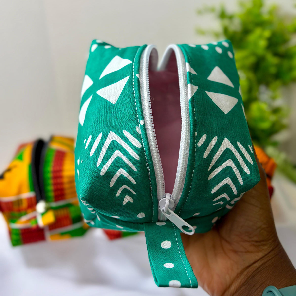 A hand holding a green and white patterned bag with a zipper, against a blurred background of a plant.