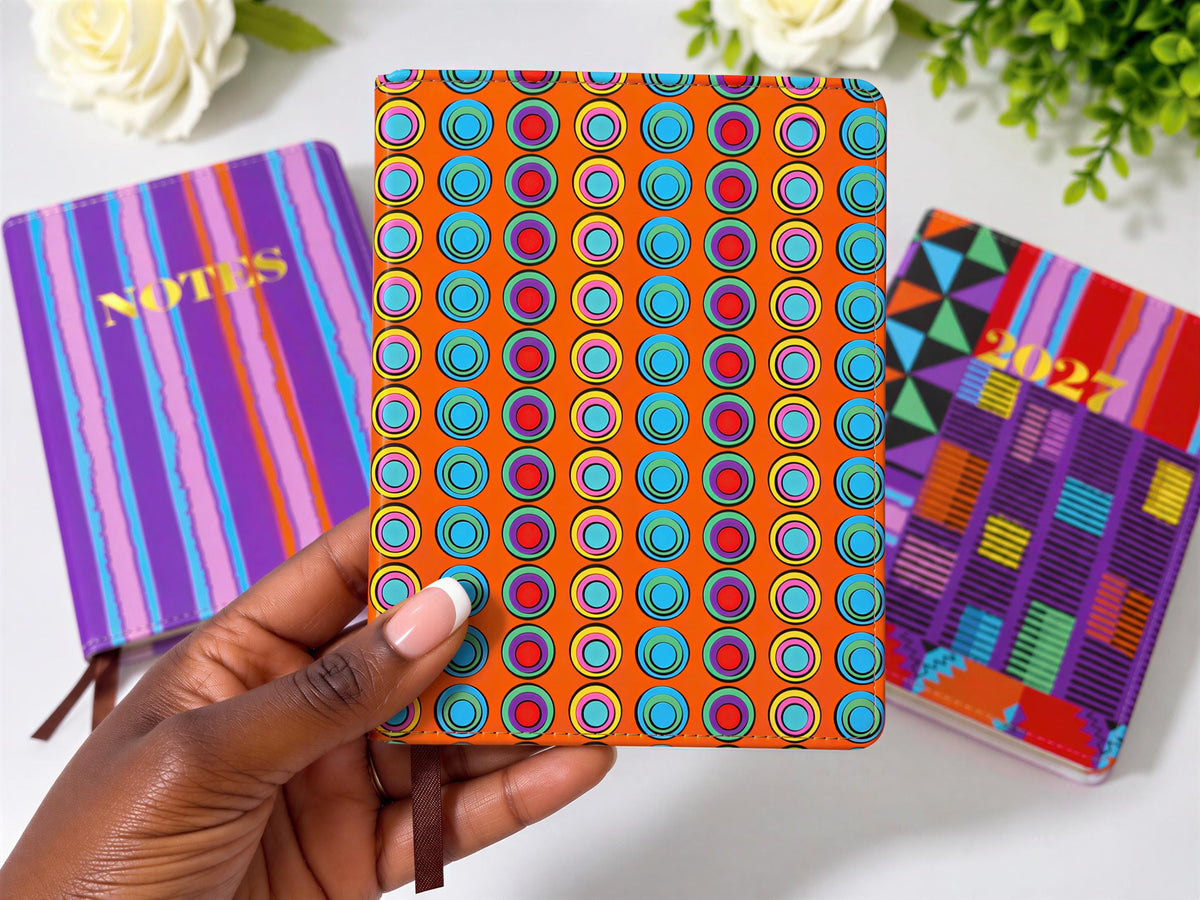 A hand holding a colorful notebook with a pattern of circles and dots, surrounded by other colorful notebooks and flowers on a white surface.