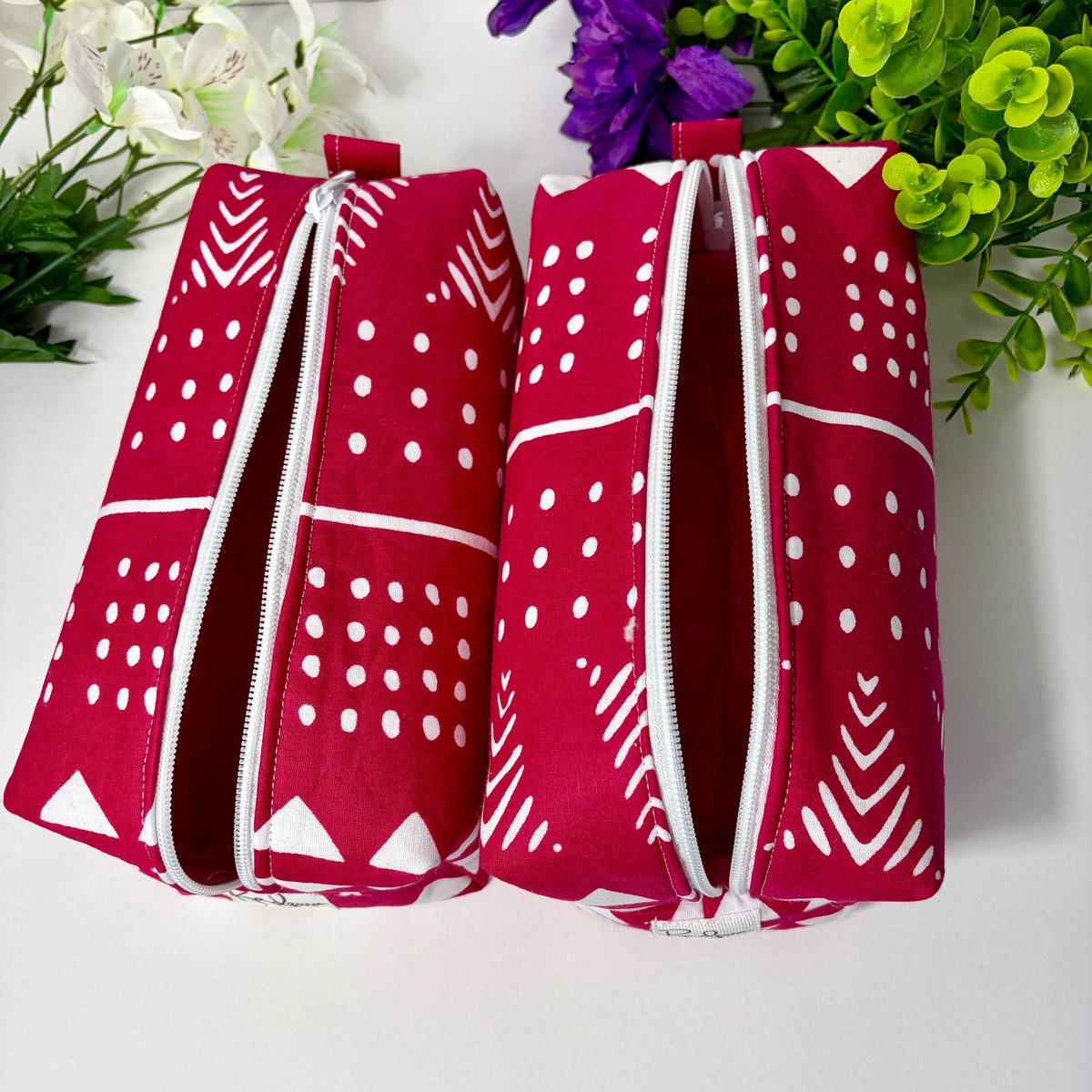 Two red and white patterned bags with zippers are placed on a white surface, surrounded by white flowers and green foliage.