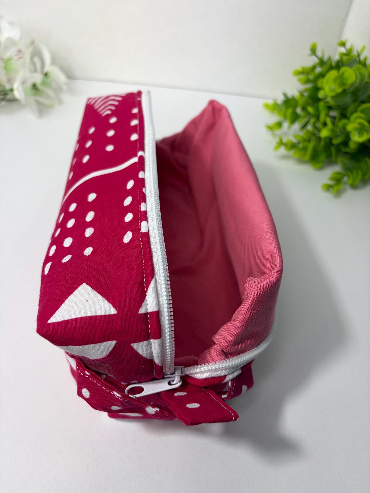 A red and white polka dot makeup bag with a pink interior is shown on a white surface, with a small green plant in the background.