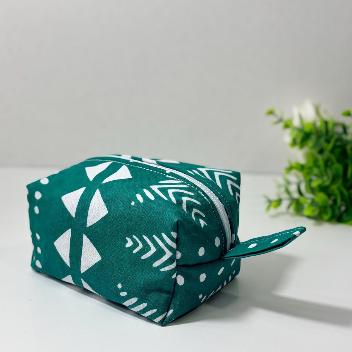 A green and white patterned bag with a zipper closure is placed on a white surface, with a green plant in the background.