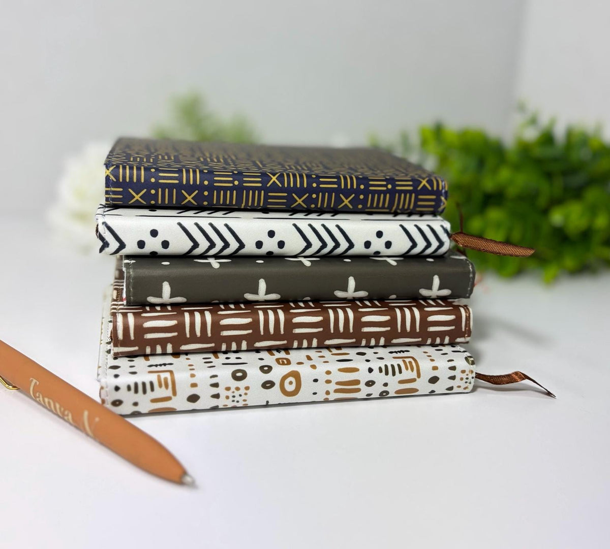 A stack of six patterned notebooks is arranged on a white surface, with a pen lying next to them.