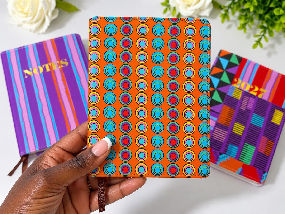 A hand holding a colorful notebook with a pattern of circles and dots, surrounded by other colorful notebooks and flowers on a white surface.