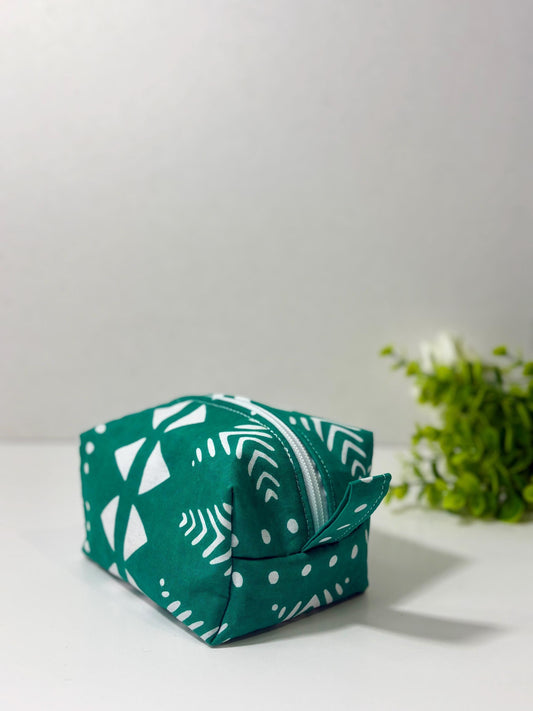 A green and white patterned bag with a zipper closure is placed on a white surface, with a small plant or greenery visible in the background.