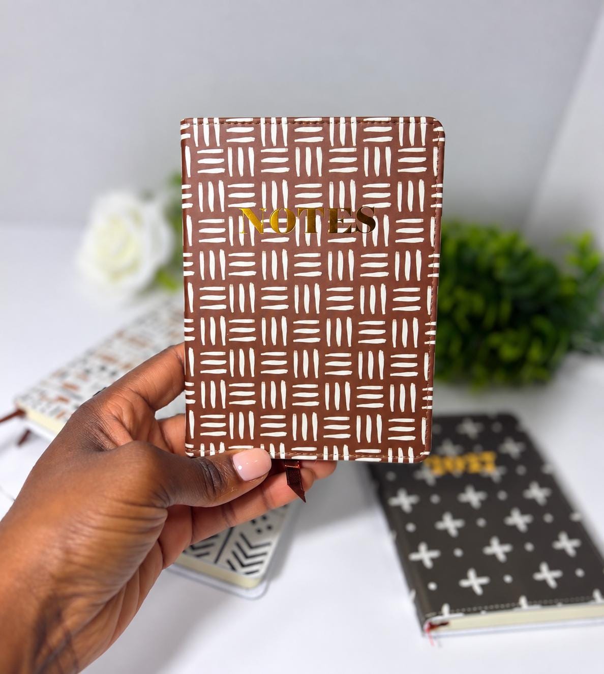 A hand holding a brown and white patterned notebook with the word "NOTES" printed on it, against a blurred background of other notebooks and a white flower.